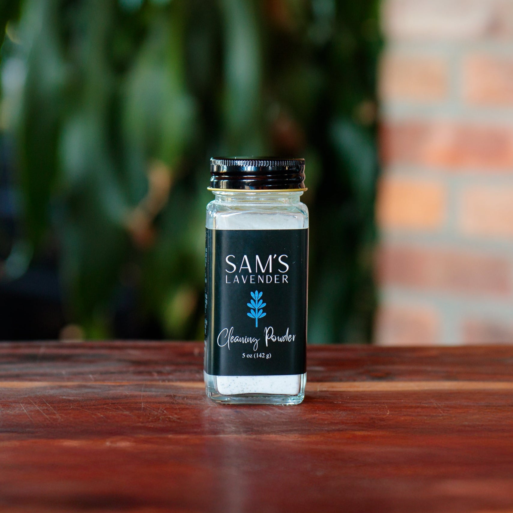 A clear glass jar labeled 'Sam's Lavender Cleaning Powder' with a black lid, on a wooden surface with foliage in the background.