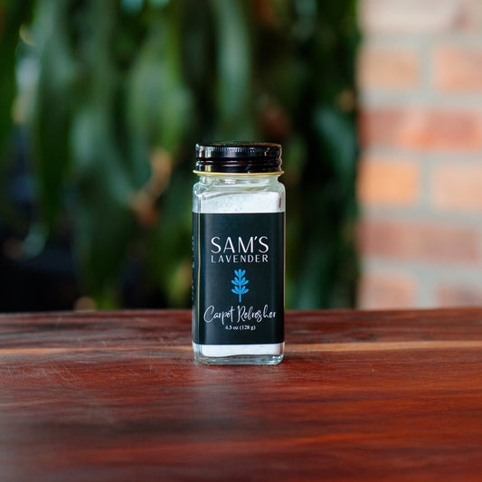 A clear glass jar of 'Sam's Lavender Carpet Refresher' with a black lid, placed on a wooden surface, with a blurred background of brick wall and greenery.