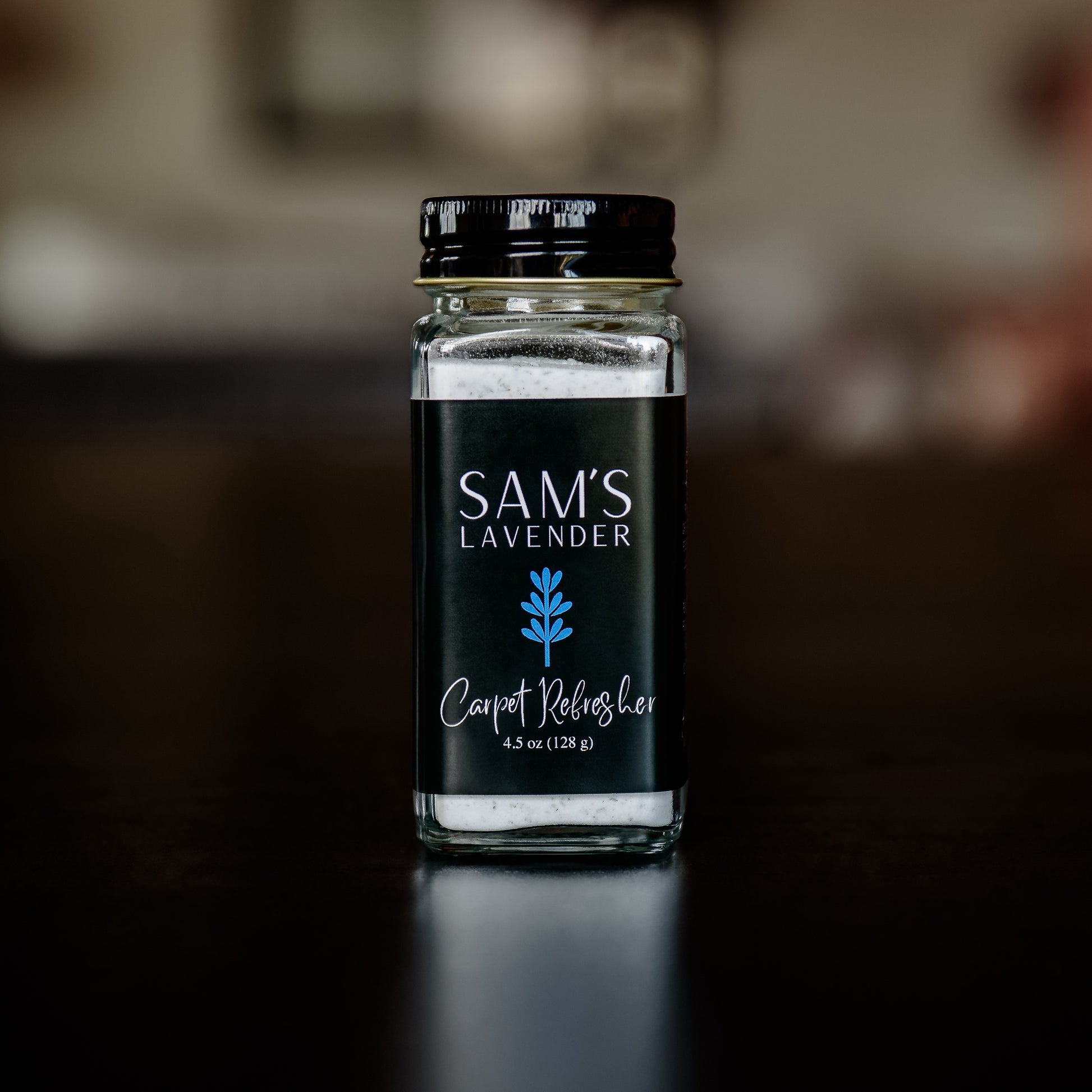 A clear glass jar of 'Sam's Lavender Carpet Refresher' with a black lid, placed on a wooden surface, with a blurred background of brick wall and greenery.