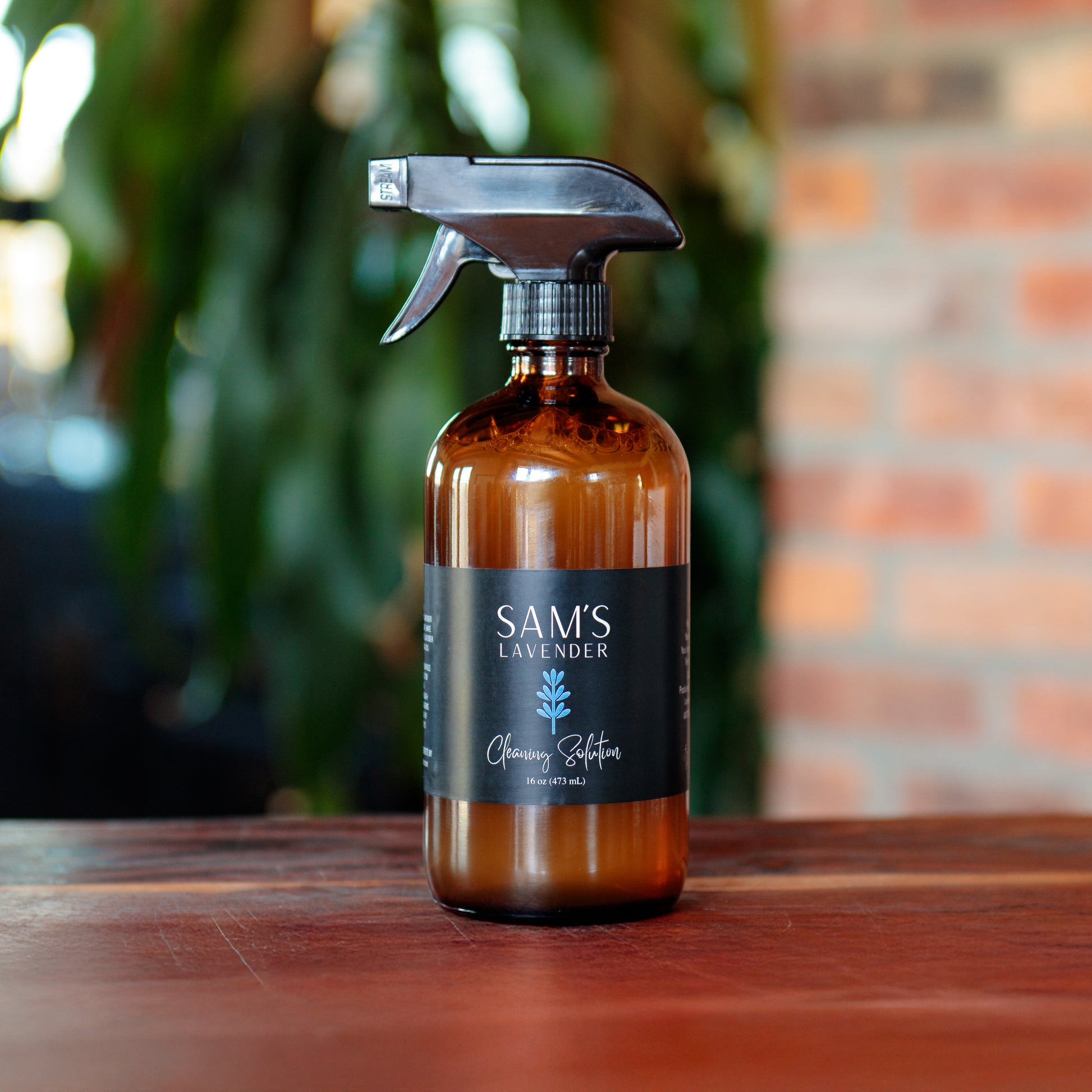 Amber glass 16oz spray bottle labeled 'Sam's Lavender Cleaning Solution' placed on a wooden surface with a blurred background featuring greenery and a brick wall.