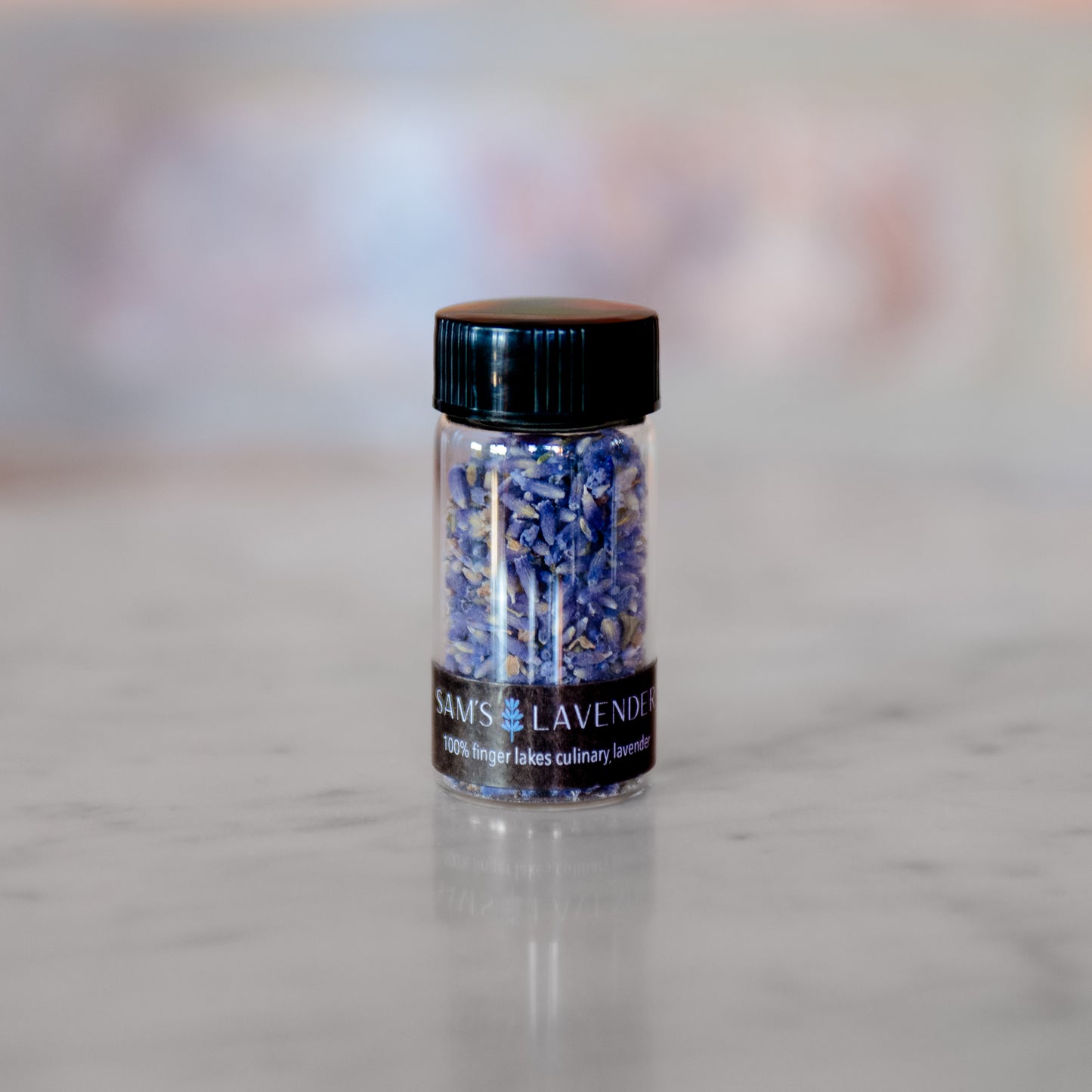 A glass jar containing culinary lavender buds on a wooden surface, with a black lid and a label that reads 'SAM'S LAVENDER 100% Finger Lakes culinary lavender'.