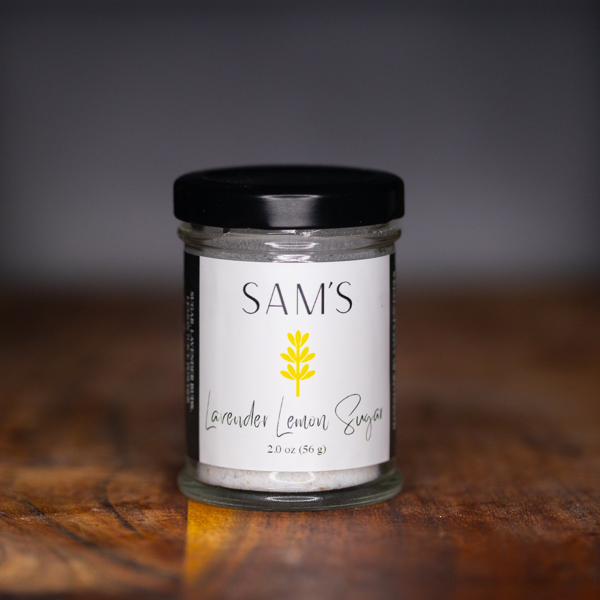 A glass jar of Sam's Lavender Lemon Sugar placed on a wooden surface with a blurred background.