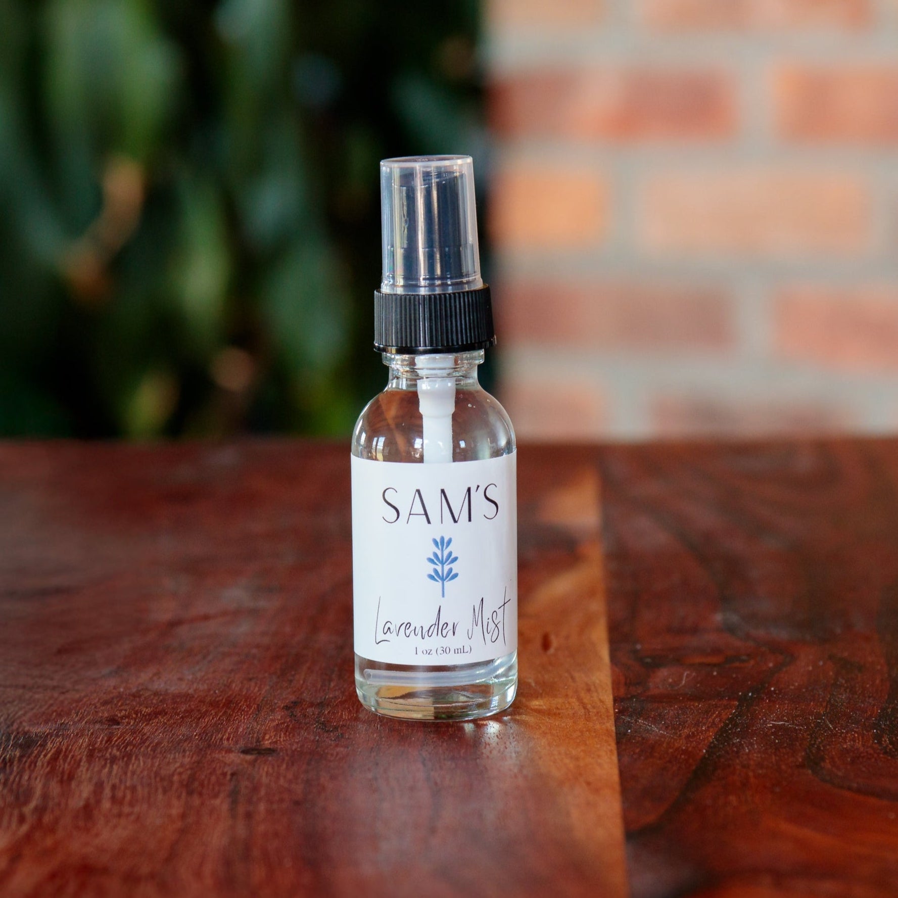A clear glass 1oz spray bottle labeled 'Sam's Lavender Mist' on a wooden surface with a blurred background.