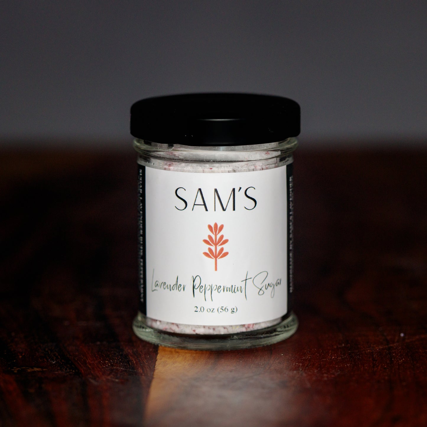 A glass jar of Sam's Lavender Peppermint Sugar on a wooden surface with a blurred background.