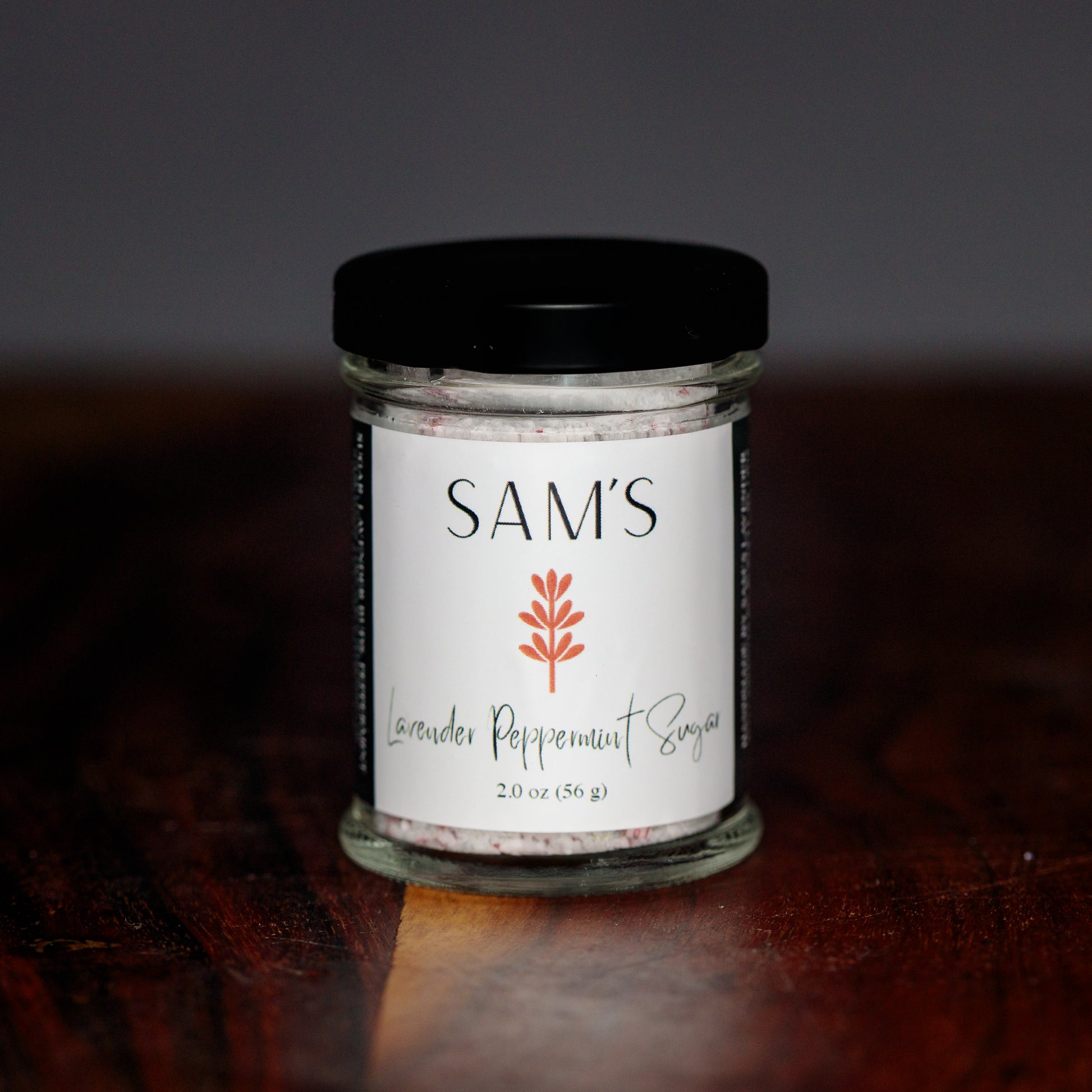 A glass jar of Sam's Lavender Peppermint Sugar on a wooden surface with a blurred background.