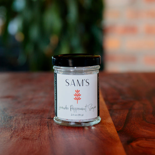 A glass jar of Sam's Lavender Peppermint Sugar on a wooden surface with a blurred background.