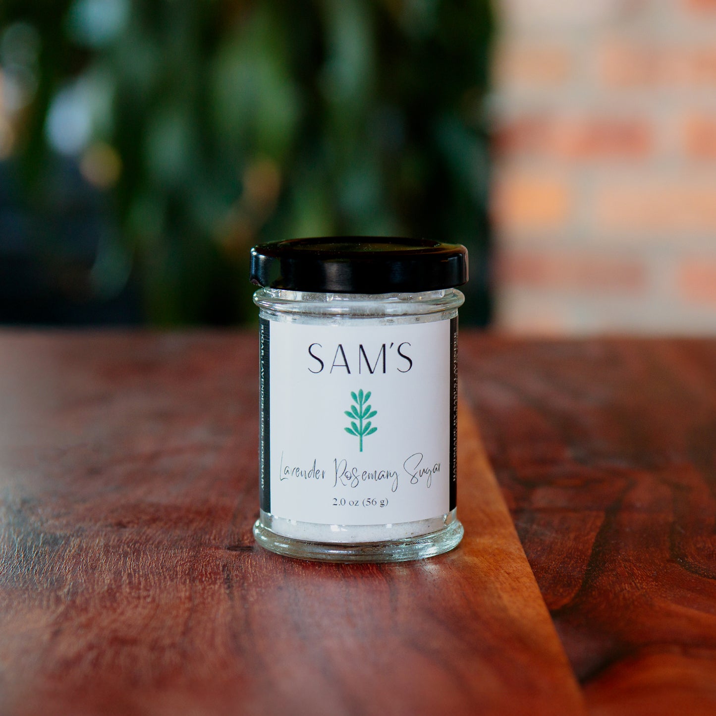 A glass jar of Sam's Lavender Rosemary Sugar placed on a wooden surface with a blurred background.
