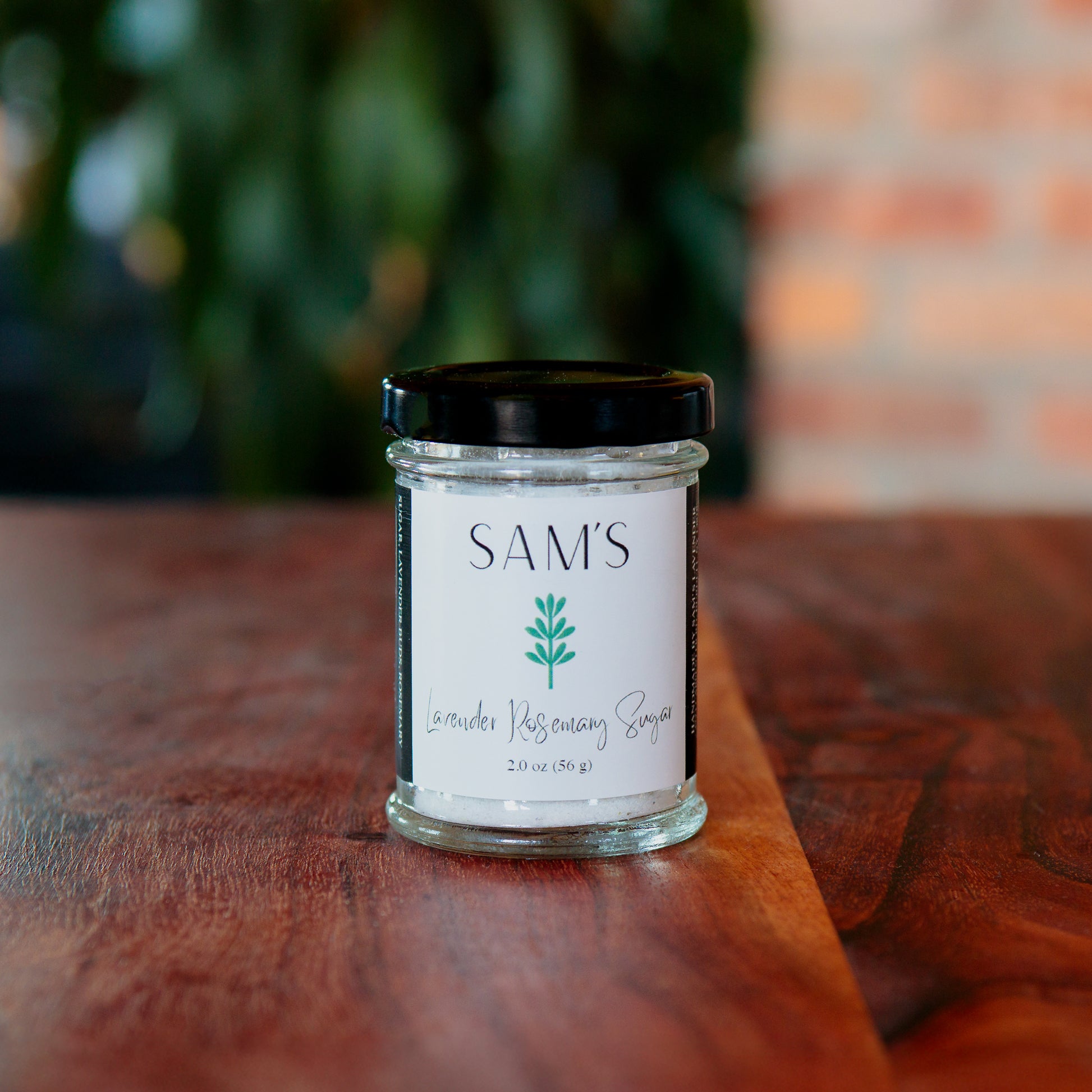A glass jar of Sam's Lavender Rosemary Sugar placed on a wooden surface with a blurred background.