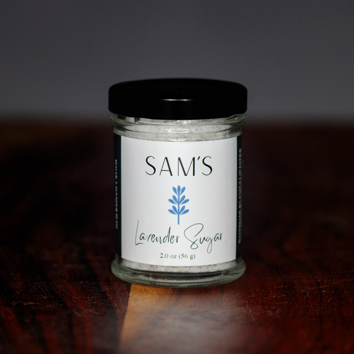 A glass jar labeled 'Sam's Lavender Sugar 2oz' containing lavender sugar, placed on a wooden surface.