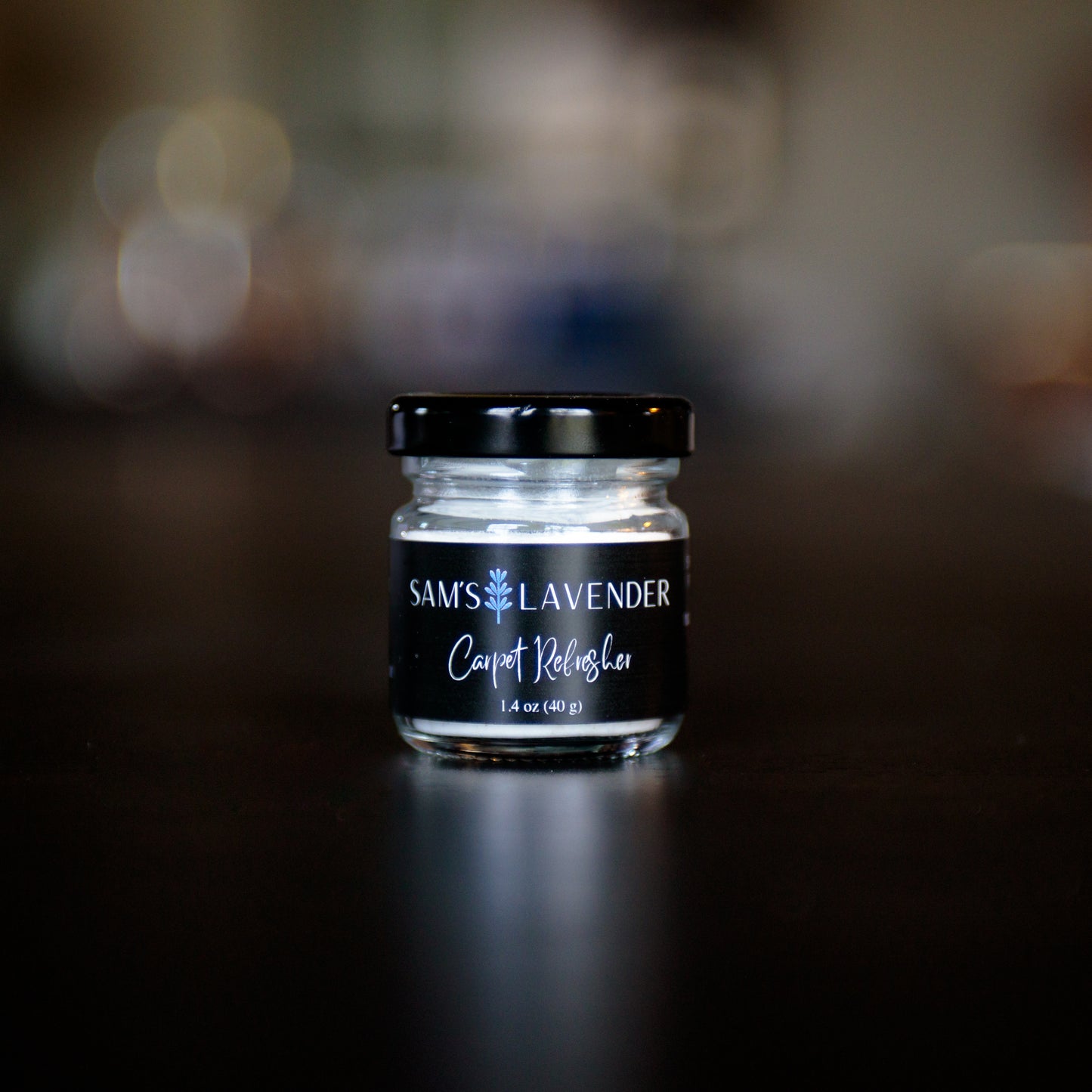 A mini clear glass jar with a black lid labeled "Sam's Lavender Carpet Refresher 1.4 oz"