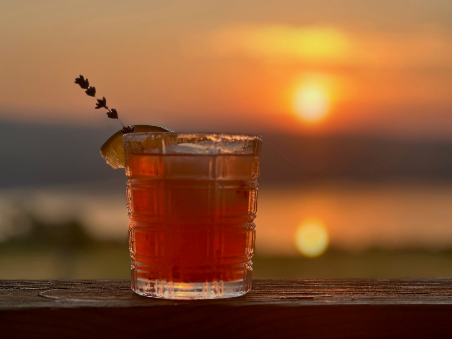 A rocks glass with a cocktail garnished with a culinary lavender stick, with a sunset in the background.
